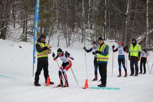 Nordic ski racers are lined up in order of their bib numbers at the UP Nordic Center. At the front of the line a racer crouches in anticipation of the her start in the pursuit race flanked by two race officials.