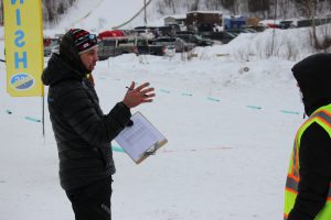 Against a backdrop of snow and a finish line flag the timing official dressed in a black hat, jacket and pants and holding a clipboard with papers explains something to a race volunteer at the UP Nordic Center.